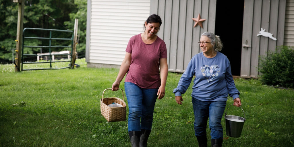 Two women are walking towards the camera. One is wearing a deep rose colored short sleeve shirt and blue denim jeans. She has dark hair tied back and is caring a woven basket. The women to the right is wearing glasses. She has long grey hair that has been pulled back she is wearing a blue long sleeve shirt with a heart image on the front. She carries a milk bucket in her hand and is wearing dark denim jeans. It is late spring and the grass and trees are green and lush.