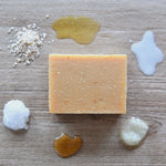 A bar of honey and oats goat milk soap is surrounding by the ingredients that go into making that soap. 