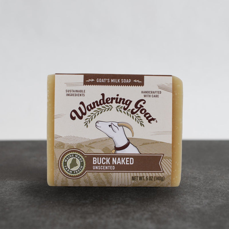 A wrapped bar of wandering goat Buck Naked goat milk soap sits on a piece of grey slate with a white background.