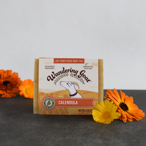 A wrapped bar of wandering goat calendula soap sits on a piece of slate with fresh calendula flowers on the lower right corner of the soap and behind the soap on the left.