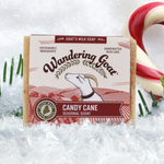 a wrapped bar of wandering goat Candy Cane goat milk soap is in a scene with snow crystals, pine needles and a candy cane.