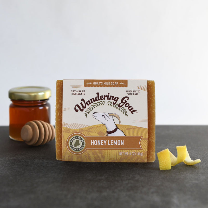 A bar of Honey Lemon goat milk soap sits on a slate board with a lemon twist in the right corner and a jar of honey in the far left background.