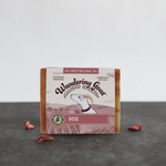 A bar of rose goat milk soap sits on a piece of slate with rose petals strewn around the base.
