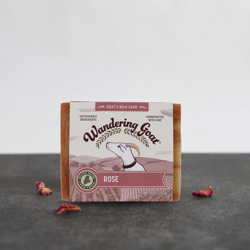 A bar of rose goat milk soap sits on a piece of slate with rose petals strewn around the base.